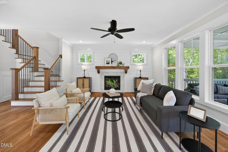 Bright living room with hardwood floors, a tiled fireplace on a shiplap wall, a staircase, and large windows.