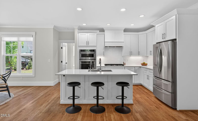 A bright kitchen with white shaker cabinets, stainless steel appliances, a center island with black stools, and hardwood floors.