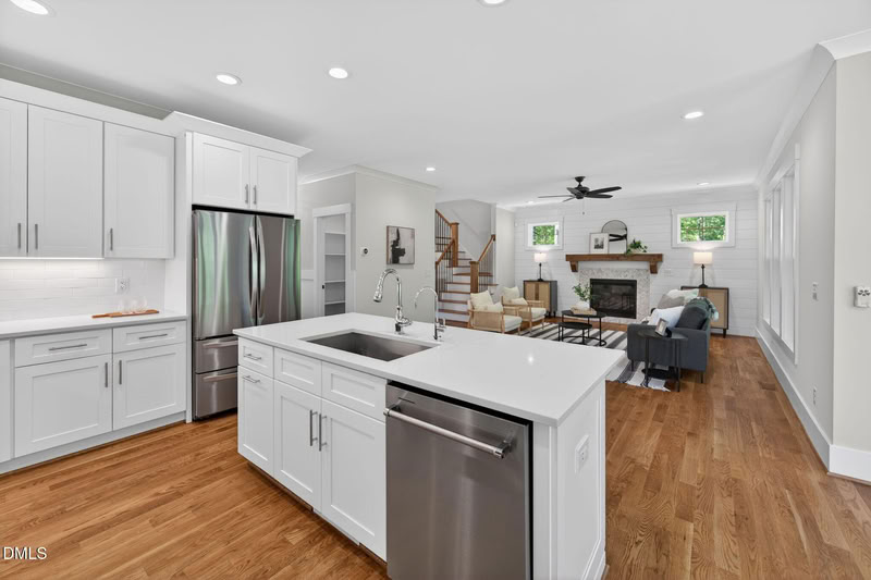Open-plan kitchen with white cabinets, a quartz island, and hardwood floors overlooking a bright living room with a fireplace.