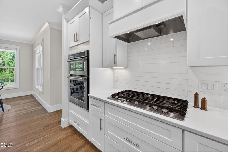 A modern kitchen with white shaker cabinets, stainless steel double ovens and gas cooktop, white quartz counters, and wood floors.