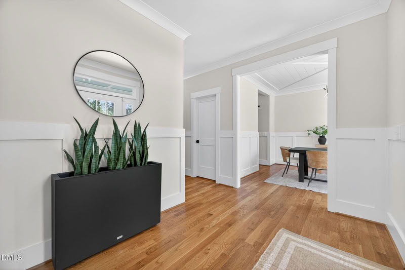 A bright hallway with hardwood floors, white wainscoting, and a round mirror. A doorway opens to a dining room in the background.