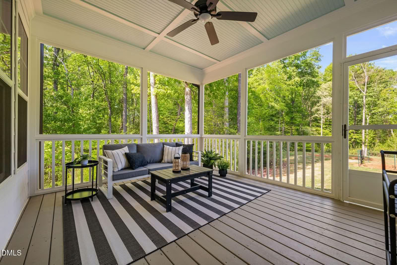 A bright, furnished screened-in porch with a ceiling fan, grey wood floors, and views of a lush green forest.