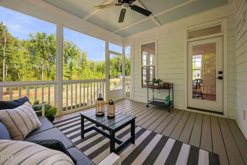 Furnished screened-in porch with wood deck, blue beadboard ceiling, ceiling fan, and large windows overlooking a wooded yard.