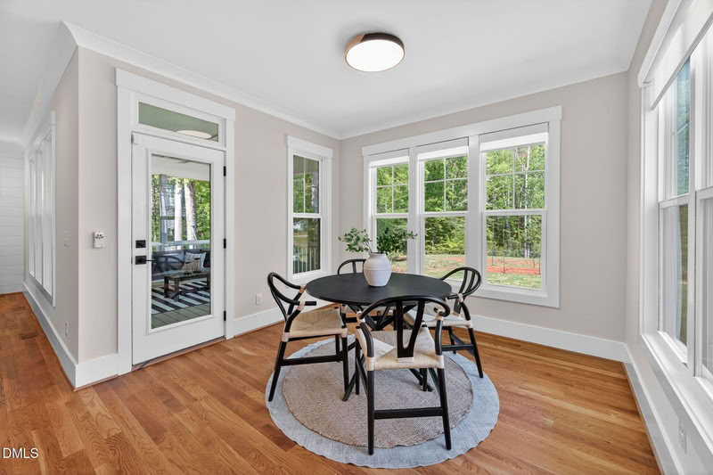 Sunlit dining nook with hardwood floors, a round black table, and large windows overlooking a leafy backyard.