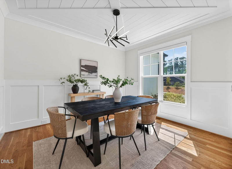 A bright dining room with a dark wood table, woven chairs, white wainscoting, hardwood floors, and a modern black chandelier.