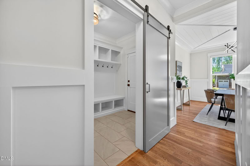 A modern sliding barn door separates a hardwood hallway from a tiled mudroom with white built-in benches and hooks.