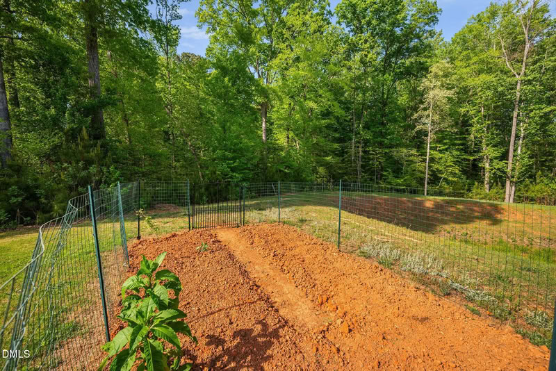 A fenced-in garden plot with tilled red soil, ready for planting, set against a backdrop of lush green trees on a sunny day.