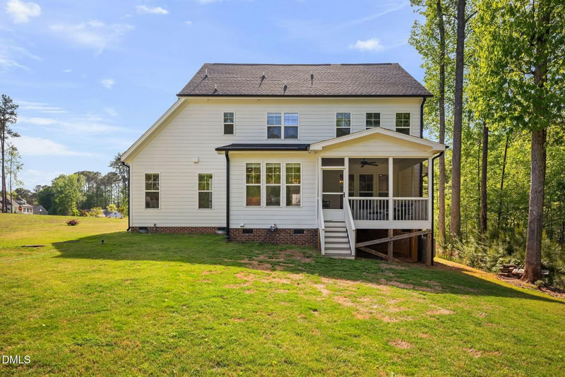 Rear view of a two-story home with light siding, a screened porch, and a large grassy backyard sloping towards a wooded area.