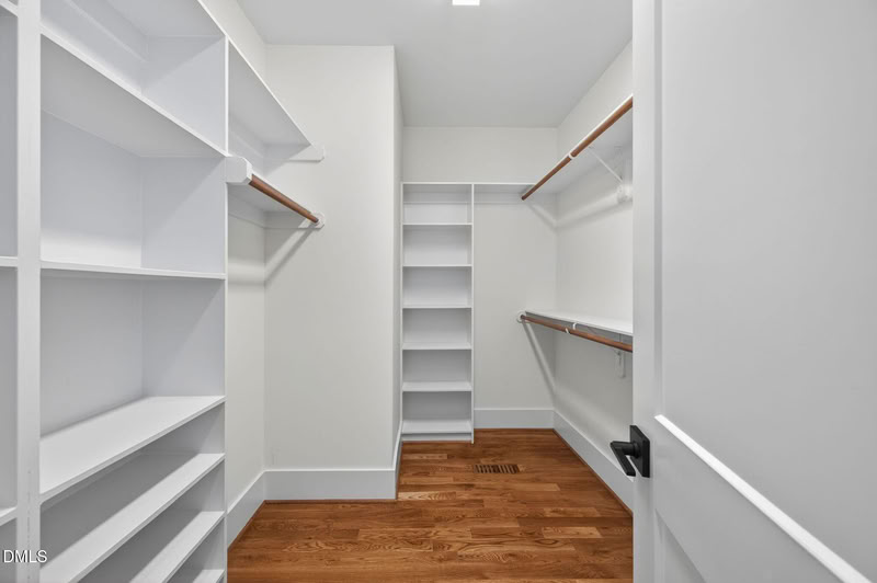 A bright, empty walk-in closet with white built-in shelving, wood hanging rods, and light hardwood floors.