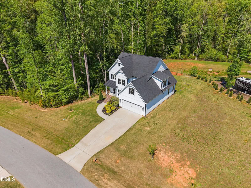 Aerial view of a two-story white house with a dark roof, long driveway, and large grassy yard backing onto a dense forest.